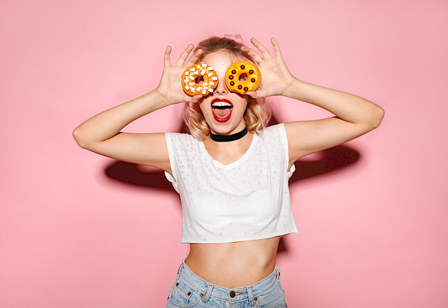 Frau hält sich lachend zwei Donuts vor die Augen – Fotobox-Spaß vor rosa Hintergrund